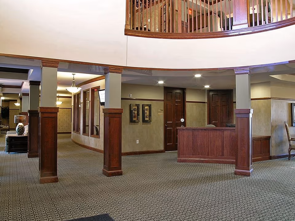 Interior view of a senior living facility lobby area with carpeted floor, wooden pillars, a reception desk, and a curved balcony railing above. The space is well-lit with ceiling lights and has a warm, inviting atmosphere.