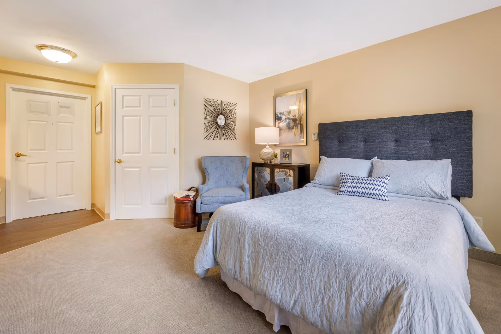 A neatly made bed with a dark gray upholstered headboard and white bedding in a bedroom. To the right of the bed is a small black nightstand with a lamp, a framed photo, and a piece of abstract wall art above it. A blue upholstered armchair sits next to the nightstand with a small round wooden side table beside it. Two white doors and a hallway are visible in the background. The room has beige walls and carpeted flooring.