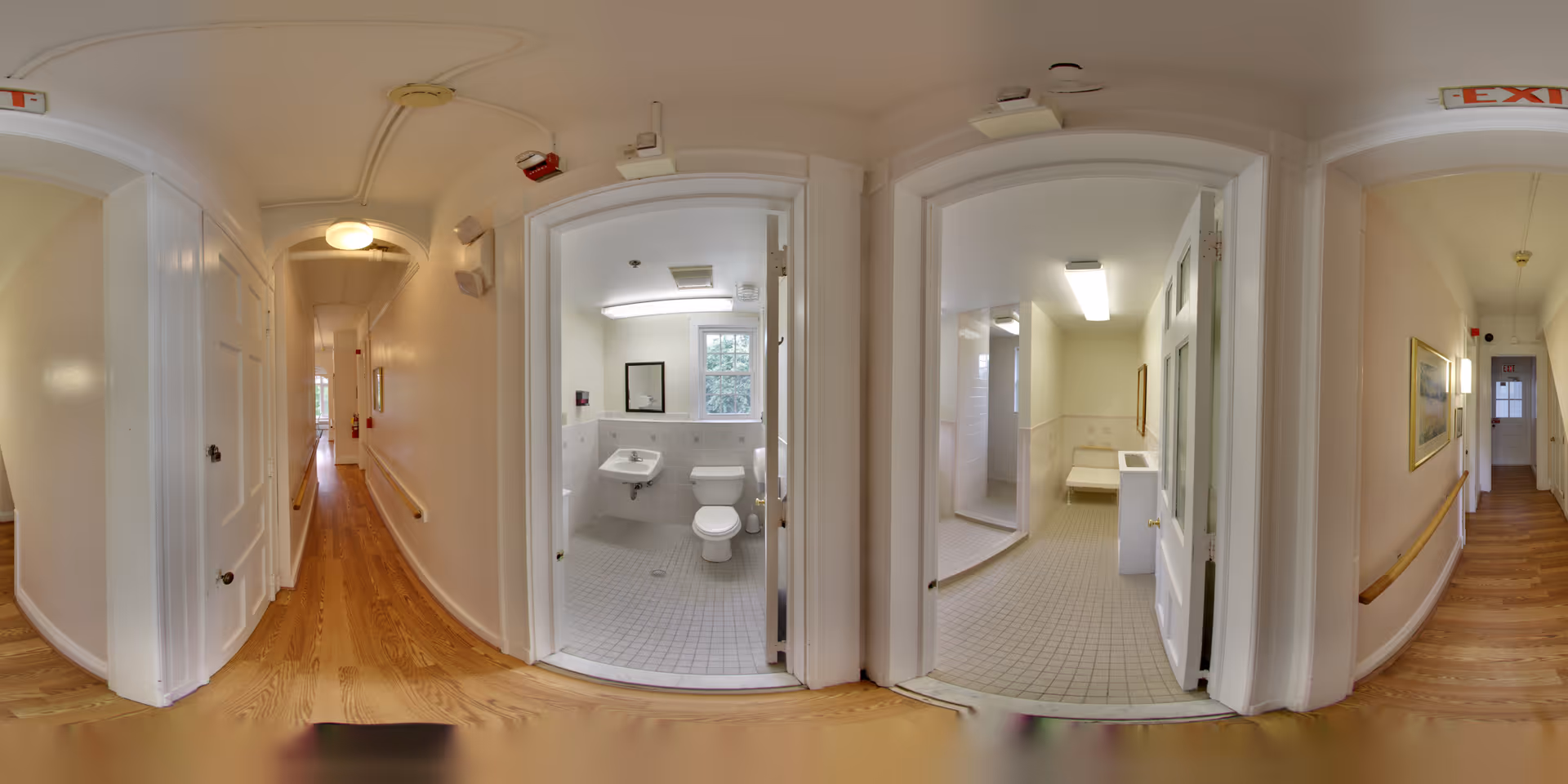 A wide-angle view of a hallway in a residential care home with wooden flooring and white walls. Two open doors reveal accessible bathrooms with tiled floors, one containing a sink, toilet, and mirror, and the other showing a shower area with a bench and sink. The hallway has handrails on both sides and exit signs above the doors.