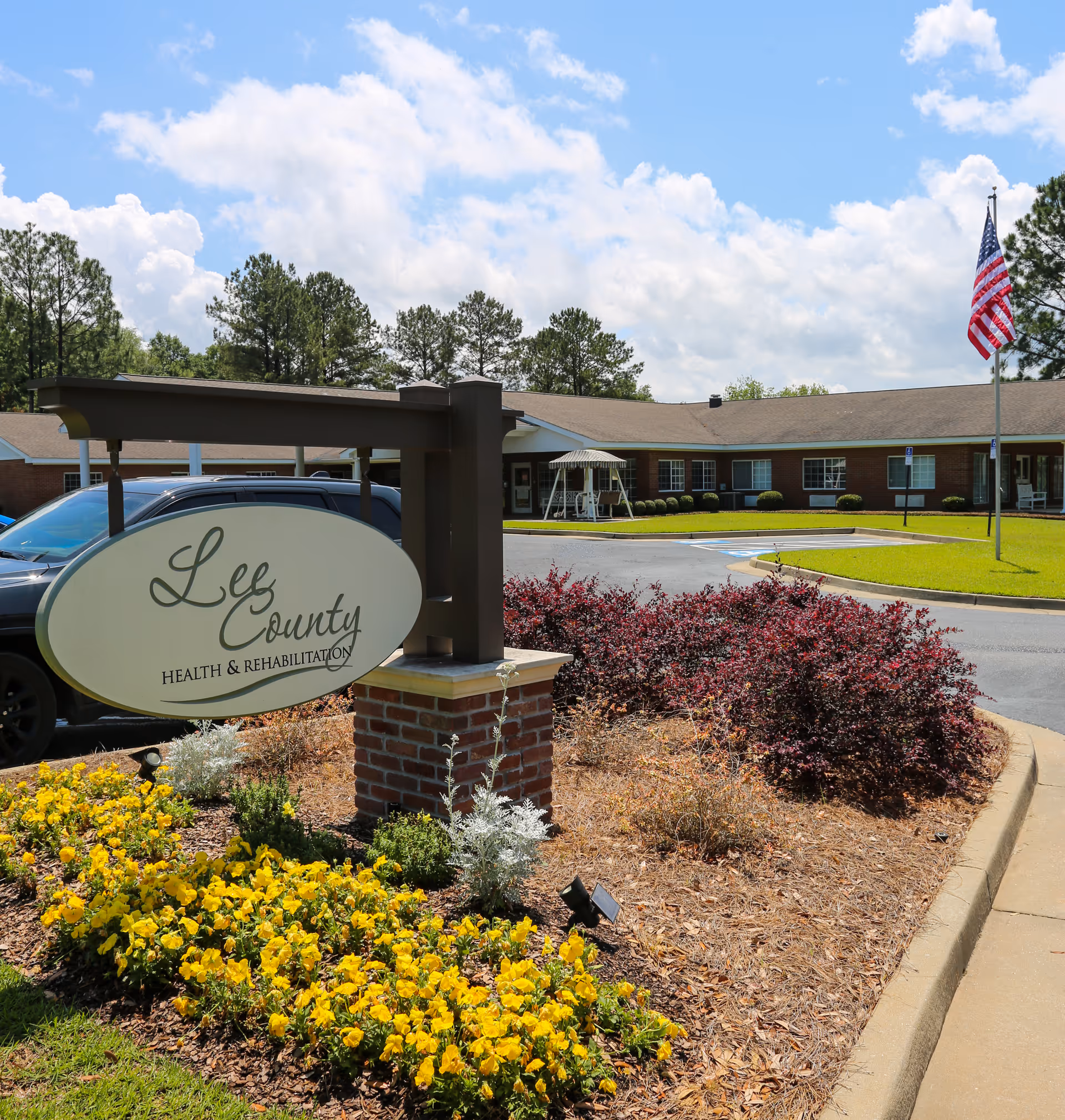 Exterior view of Lee County Health & Rehabilitation facility with a sign in the foreground surrounded by yellow flowers and landscaping. The building is a single-story brick structure with a covered entrance, an American flag on a flagpole, and a clear blue sky with some clouds.