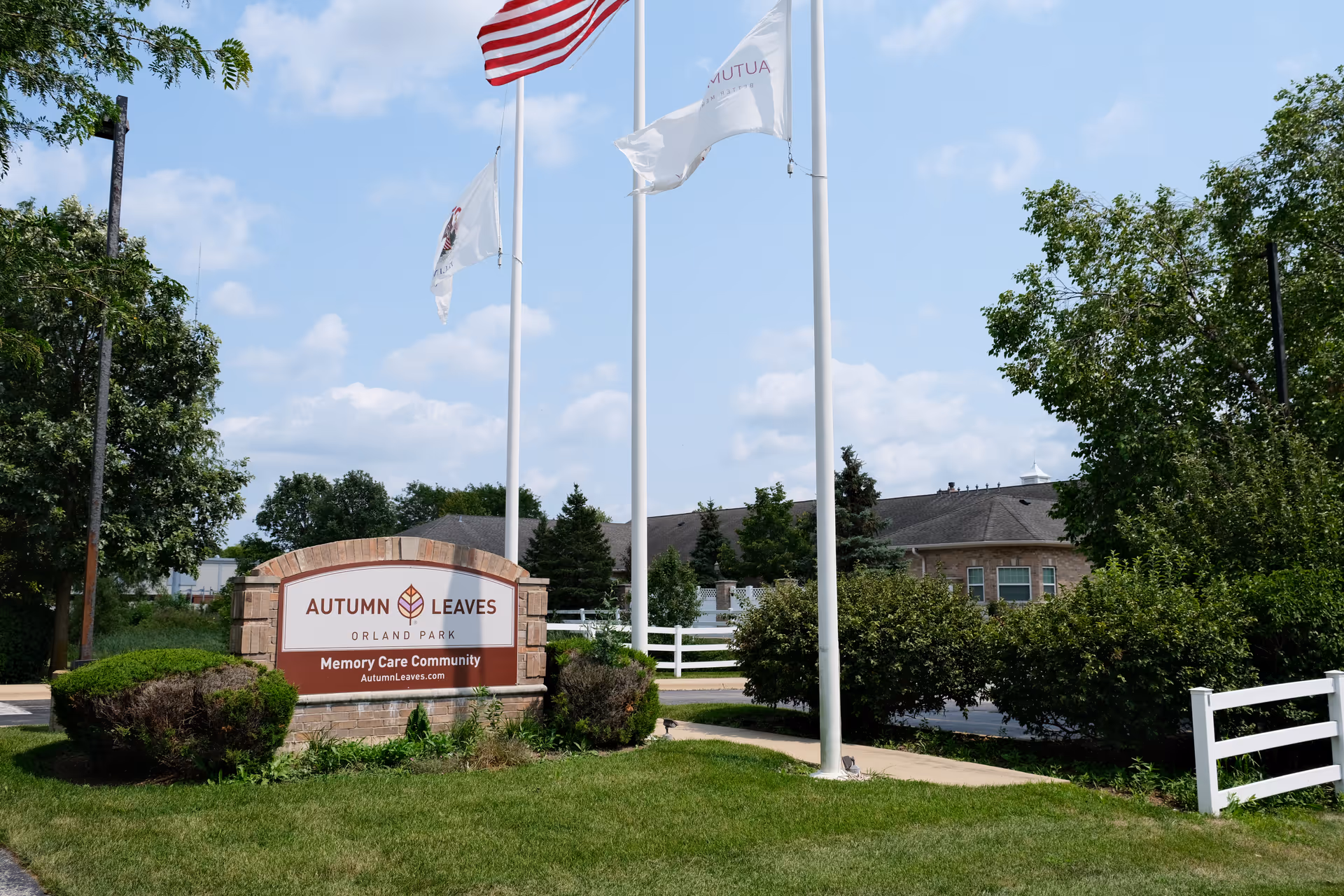 Outdoor view of the entrance sign for Autumn Leaves of Orland Park Memory Care Community, surrounded by green bushes and trees, with three flagpoles flying flags against a partly cloudy sky.