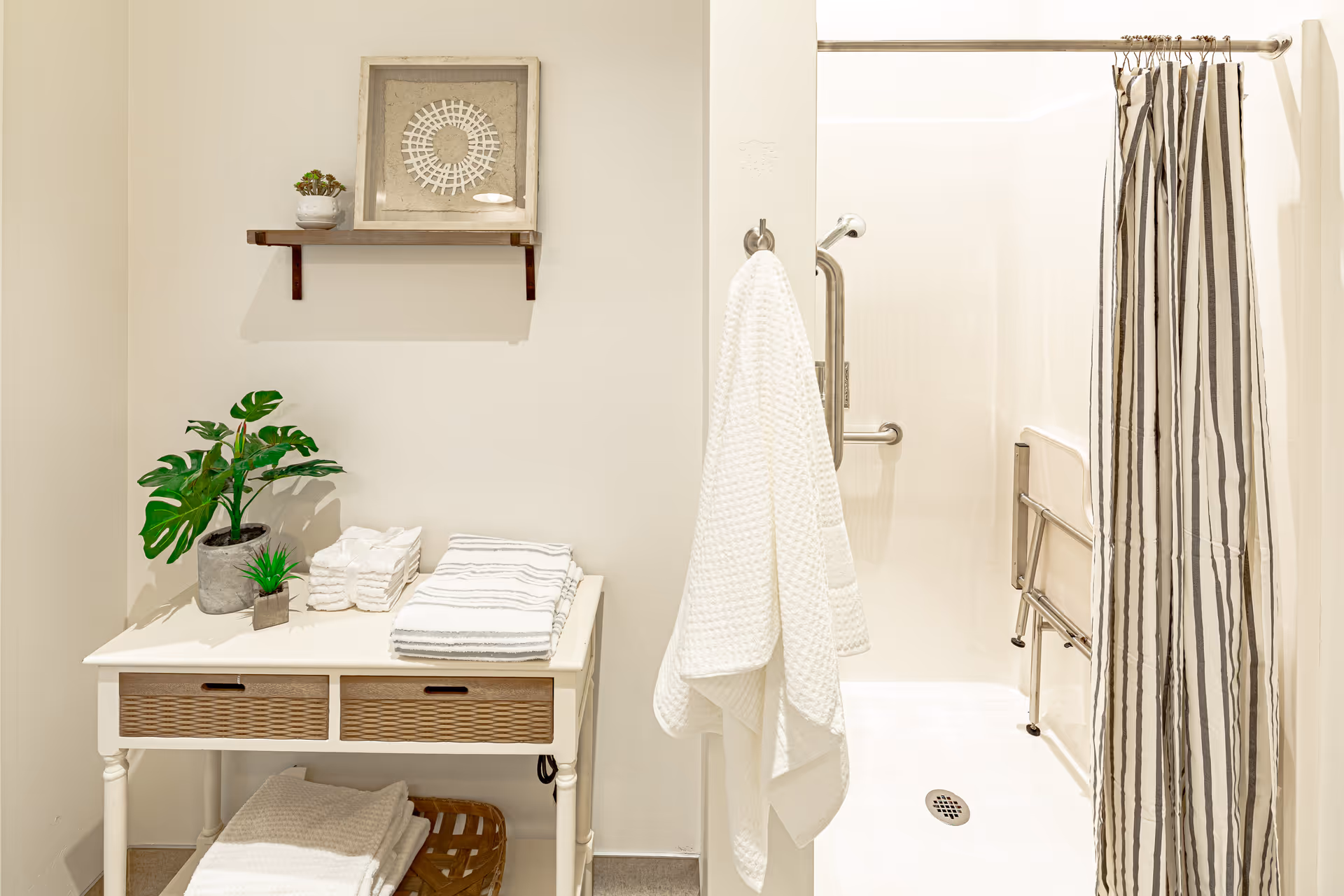 Well-lit accessible bathroom with a walk-in shower and striped curtain, grab bars, a hanging towel, and a table holding folded towels and potted plants.