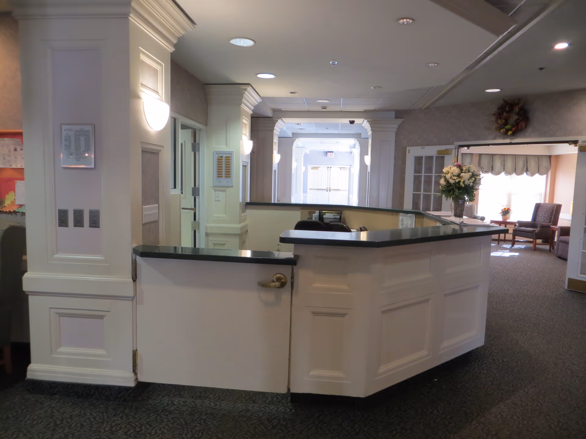 Interior view of a senior living facility reception area with a white paneled front desk featuring a dark countertop. The area is well-lit with wall sconces and recessed ceiling lights. In the background, there is a hallway leading to double doors and a sitting area with chairs, a table, and a large window with curtains. A floral arrangement is placed on the reception desk.
