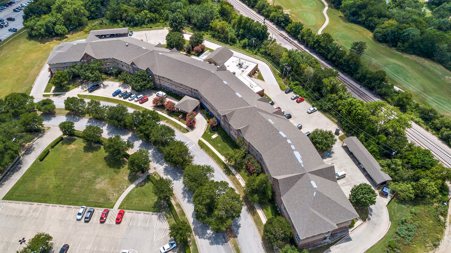 Aerial view of Morada Meadow Lakes senior living facility showing a large, curved building surrounded by trees, parking lots with cars, and adjacent green spaces including a golf course and railroad tracks.