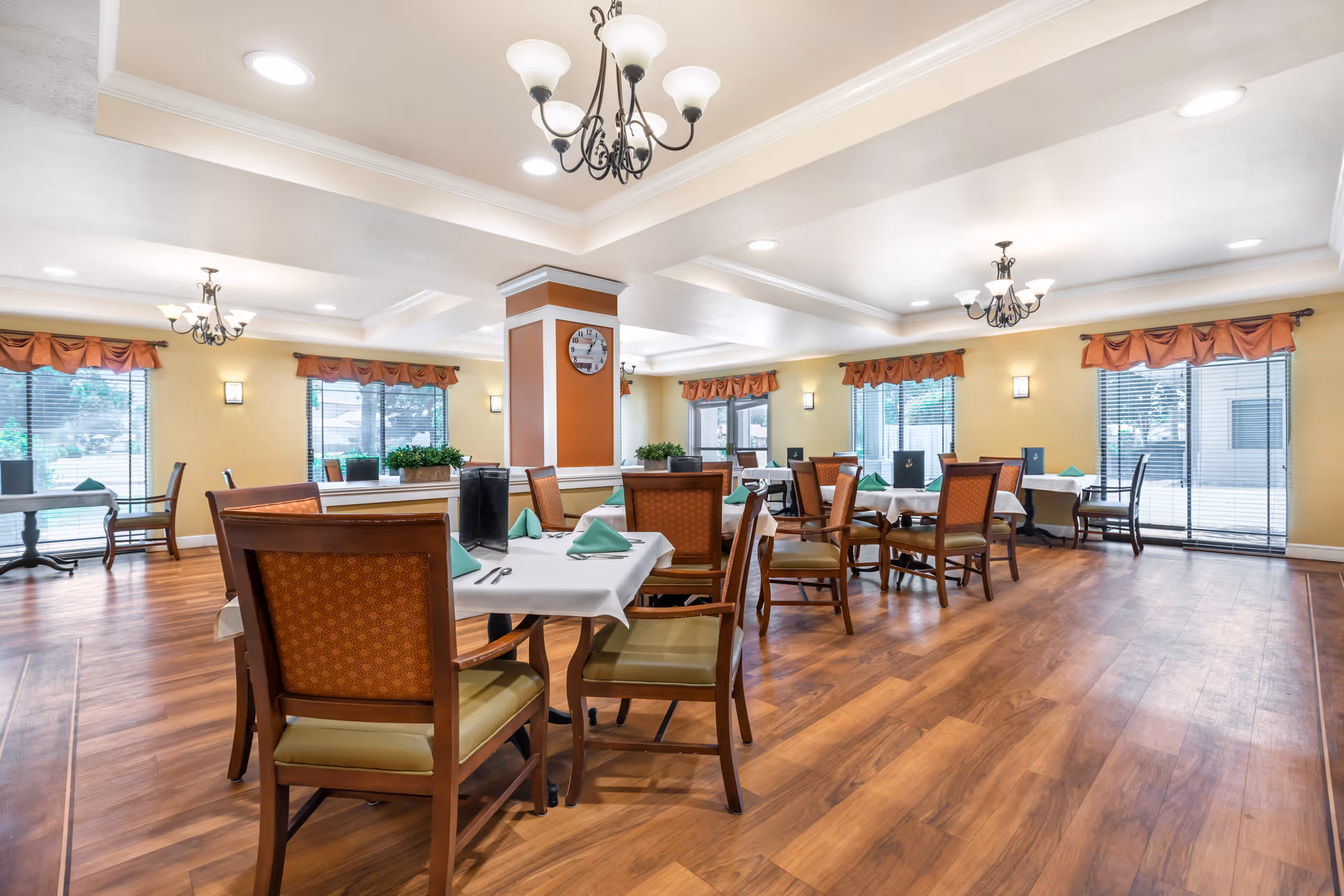 Spacious dining room with multiple set tables and wooden chairs, chandeliers, and large windows.