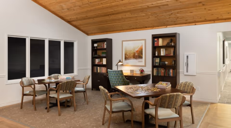 A cozy room with two wooden tables surrounded by cushioned chairs. One table has a partially completed jigsaw puzzle and a puzzle box on it. The other table has board games and cards. The room has a high wooden ceiling, two tall bookshelves filled with books, a desk with a lamp and a framed picture above it, and a window with closed blinds. There is a hallway visible to the right.