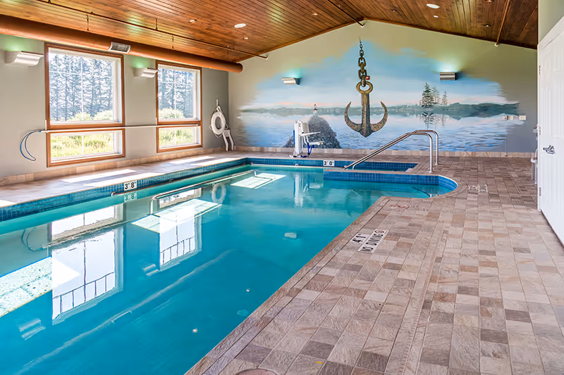 Indoor swimming pool with clear blue water, surrounded by tiled flooring and large windows letting in natural light. The ceiling is wooden with exposed beams. A mural of a large anchor hanging over a calm lake scene is painted on the far wall. Pool safety equipment is visible near the windows.