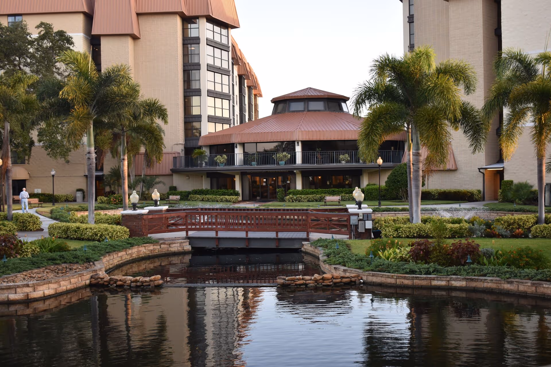 View of a senior living facility building with a pond and a small wooden bridge in the foreground, surrounded by palm trees and landscaped gardens.