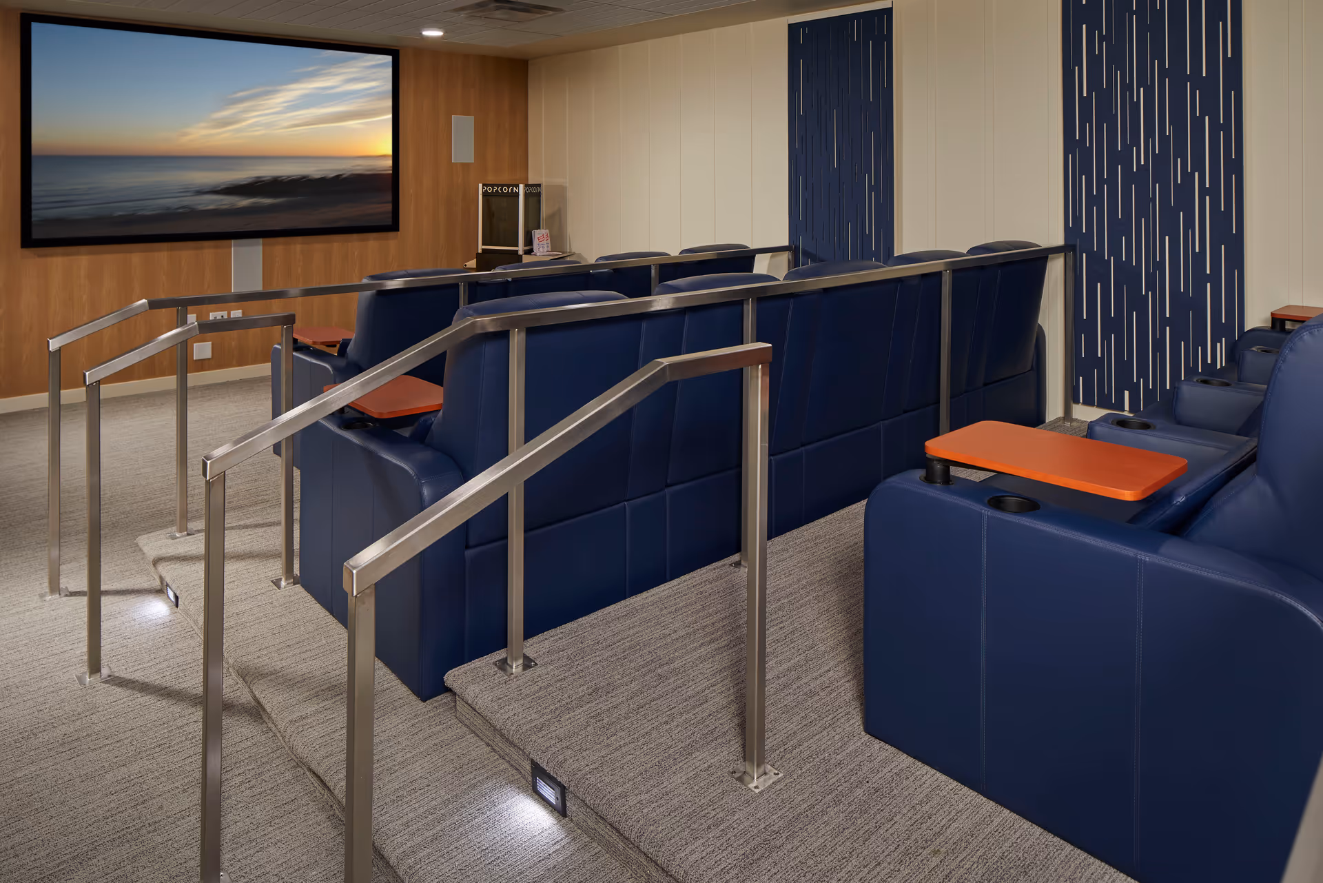 Tiered screening room with blue recliner seats, orange tray tables, metal handrails, and a large wall-mounted screen showing a seaside sunset.