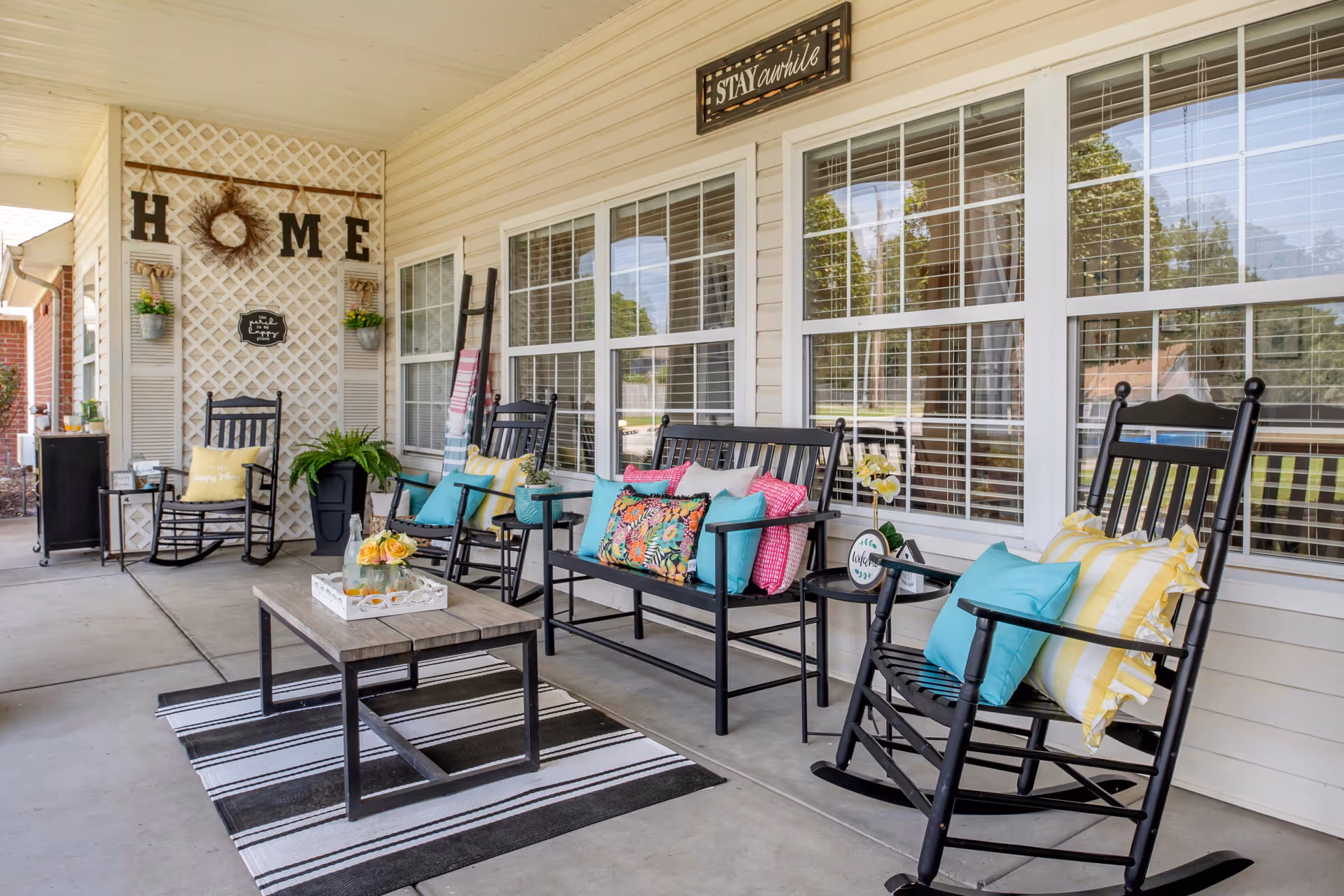 A cozy outdoor porch area with black rocking chairs and a bench adorned with colorful cushions. A wooden coffee table with a tray holding a bottle and flowers sits on a striped rug. The porch wall features a decorative lattice with the word 'HOME' and small potted plants, along with a sign that says 'STAY awhile'. Large windows with white blinds line the wall behind the seating.