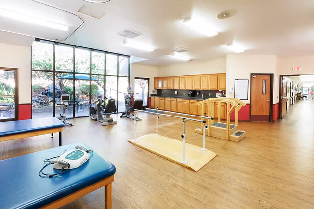 A spacious rehabilitation room with wooden flooring, featuring parallel bars for walking exercises, two blue padded therapy tables, exercise bikes near large windows overlooking an outdoor garden, and wooden cabinets along one wall. The room is well-lit with ceiling lights and has an exit door leading to a hallway.