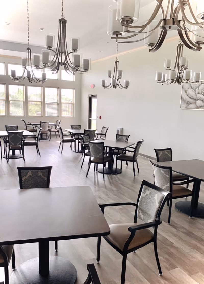A bright dining room with multiple square tables and cushioned chairs arranged neatly. The room features large windows with blinds, light-colored walls, and several modern chandeliers hanging from the ceiling. The floor is covered with wood-like flooring, and there is a door visible in the background.