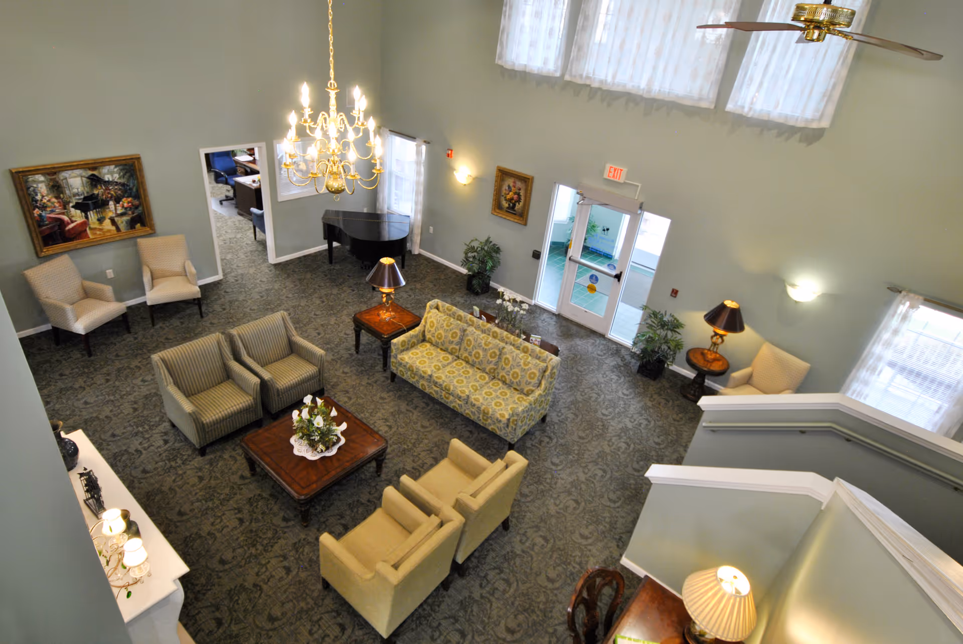 A spacious living room area in a senior living facility with multiple armchairs and sofas arranged around a central wooden coffee table. The room features a chandelier, a grand piano in the corner, framed paintings on the walls, several table lamps, and large windows with sheer curtains allowing natural light to enter. There is also a staircase visible on the right side and an exit door with glass panels.