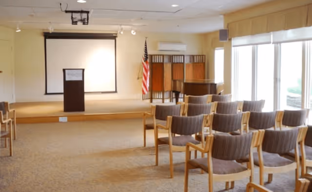 A small conference or meeting room with rows of wooden chairs facing a podium and a projection screen. An American flag stands next to the podium, and large windows on the right side let in natural light. The room has beige walls and carpeted flooring.