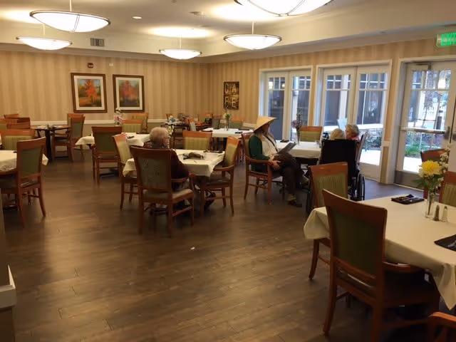 A dining room in an assisted living facility with several tables covered with white tablecloths and green cushioned chairs. A few elderly residents are seated at the tables, some in wheelchairs, near large windows and a glass door that leads outside. The room has wooden flooring, striped wallpaper, and framed artwork on the walls.