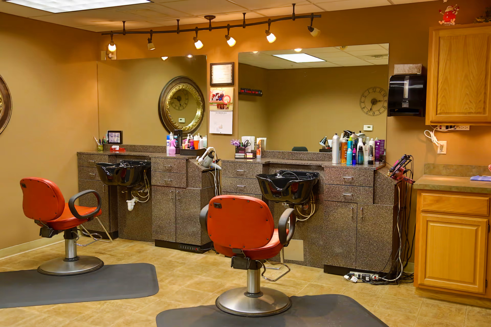 A small salon area with two red styling chairs, wash basins, mirrors, and countertops stocked with hair products.