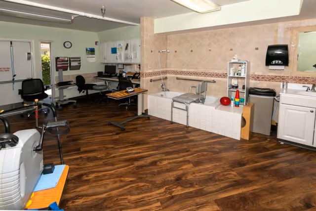 Spacious therapy room with exercise machines, a tiled therapy bathtub with a transfer bench, desks and storage cabinets.
