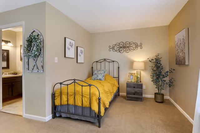A cozy bedroom with a single black metal bed frame dressed in a yellow comforter and a decorative pillow. Next to the bed is a dark wooden nightstand with a lamp, a small plant, and a picture frame. The walls are beige with decorative wall art above the bed and on the adjacent walls. A potted plant is placed in the corner near the nightstand. To the left, there is an open doorway leading to a bathroom with a sink and mirror visible.