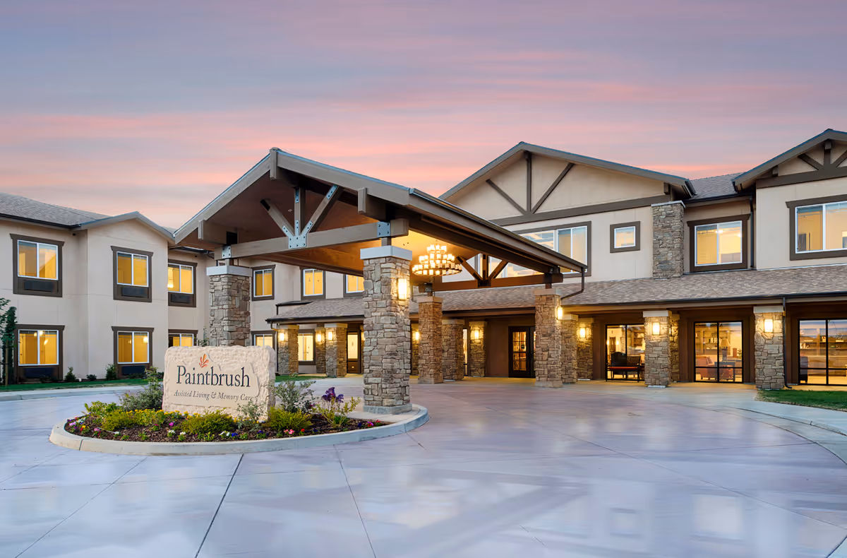 Exterior view of Paintbrush Assisted Living facility at dusk with a covered entrance supported by stone pillars, illuminated lights, and a landscaped roundabout featuring a stone sign with the facility name.