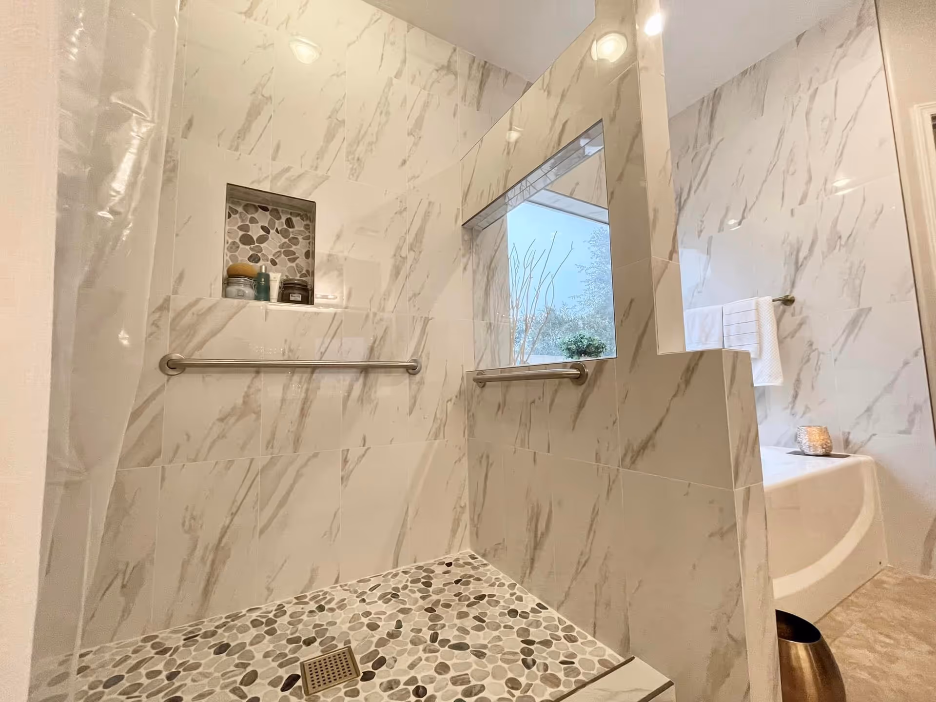 Walk-in shower with pebble-tile floor, marble-look wall tiles, grab bars, a recessed shelf, and a window opening to a bathtub area.
