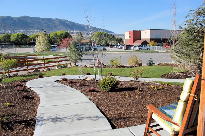 A curved concrete walkway winds through a landscaped garden toward a parking lot and a two-story building with mountains in the background, while a cushioned wooden chair sits in the foreground.