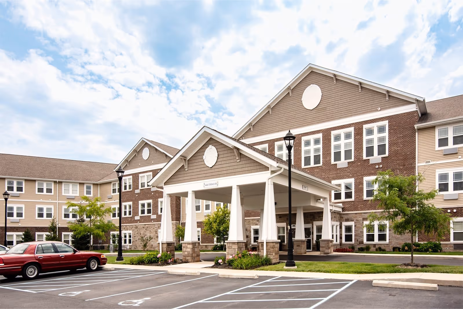 Exterior view of Woodland Terrace of New Palestine, showing a large multi-story building with a covered entrance supported by white columns. There are several windows, a few trees, street lamps, and a parking lot with marked spaces, including handicapped spots. A red car is parked in the lot under a partly cloudy sky.