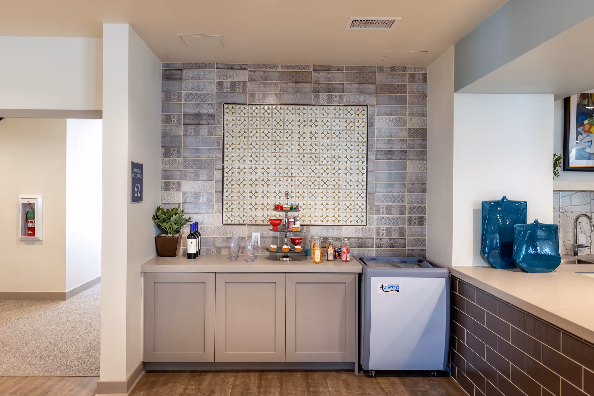 A countertop area with a decorative tiled backsplash featuring a geometric pattern. On the counter are a potted plant, two bottles of wine, several clear glasses, a tiered tray with snacks, and some small condiment bottles. To the right of the counter is a small white refrigerator with the brand name Avanti. The surrounding walls are light-colored, and there are two large blue decorative vases on an adjacent counter.