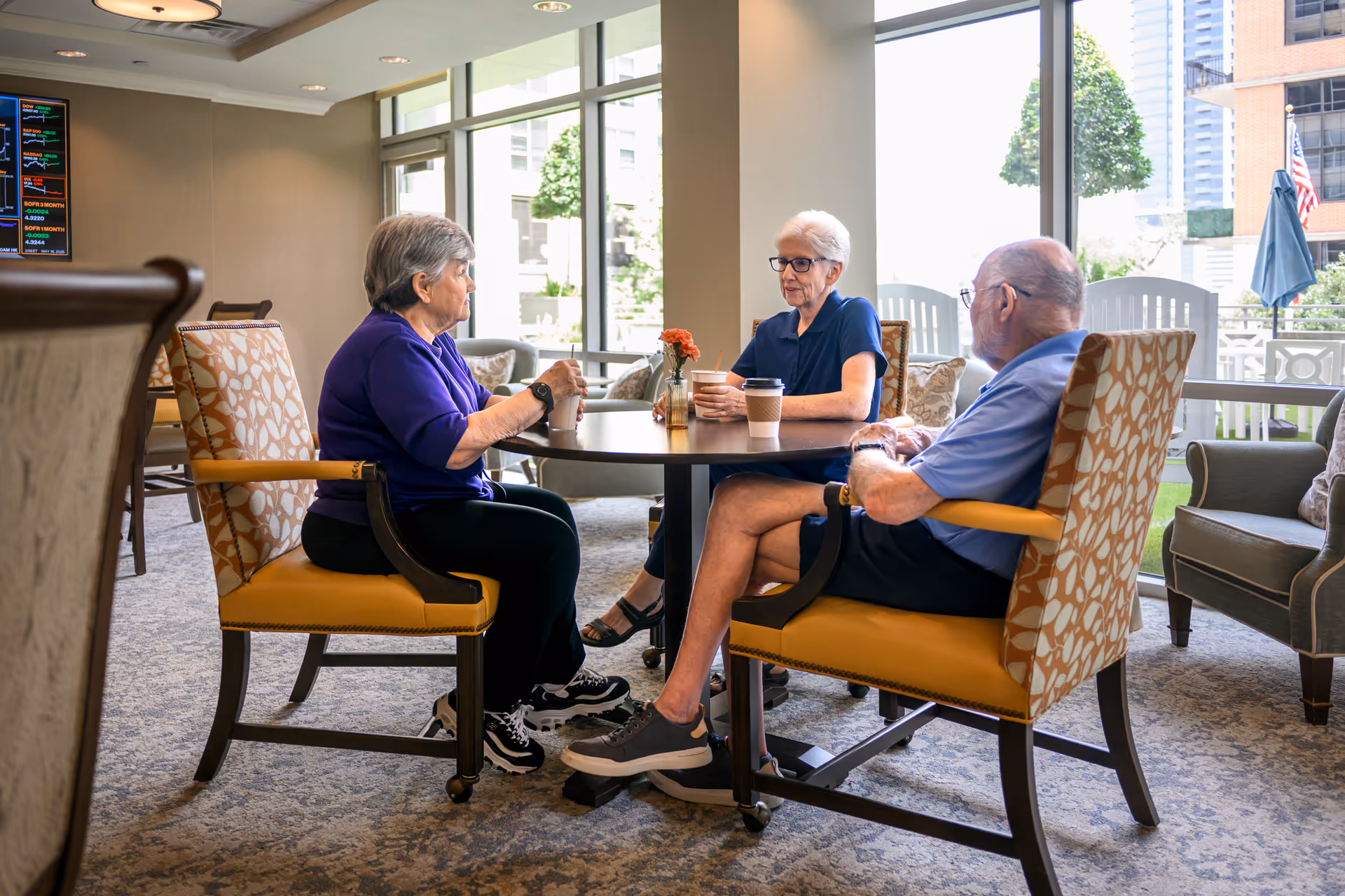 Three elderly people sitting around a round table in a well-lit common area with large windows, holding coffee cups and engaging in conversation. The room has patterned chairs, carpeted floor, and a digital display on the wall.