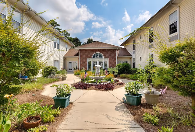 Outdoor courtyard area at Lakeview Estates - Memory Care Community featuring a central fountain surrounded by plants and small American flags, with two beige buildings on either side under a partly cloudy blue sky.