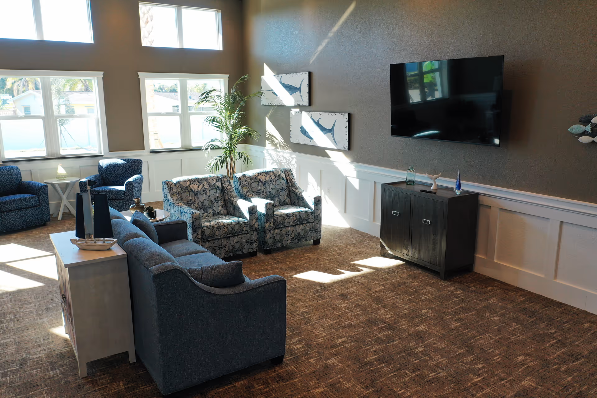 A bright living room with large windows letting in natural light. The room features a blue sofa, two floral armchairs, and two blue armchairs arranged around a small table. A flat-screen TV is mounted on the wall above a dark wooden cabinet. There are two framed fish artworks on the wall and a tall potted plant near the windows.