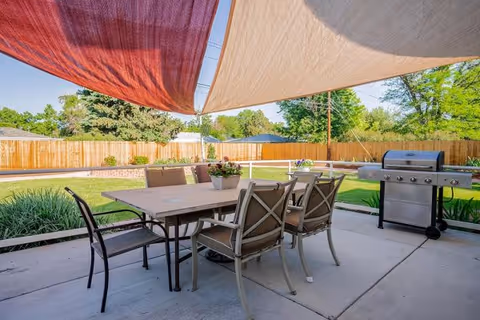Outdoor patio area with a dining table and six chairs under two large shade sails. There is a barbecue grill to the right and a wooden fence enclosing a grassy yard with trees and shrubs in the background.