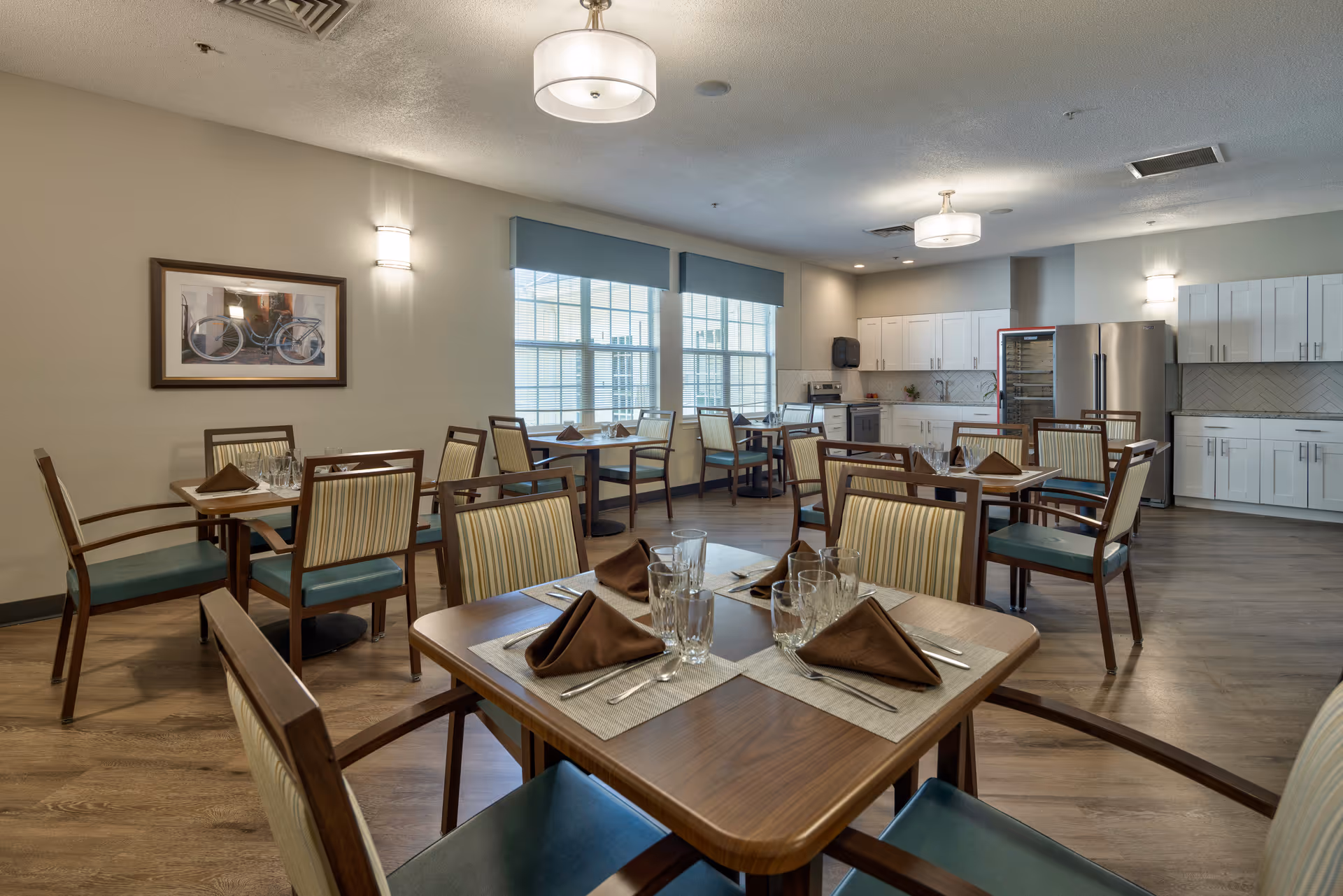 A dining room with several wooden tables and chairs arranged neatly. Each table is set with brown folded napkins, glasses, and silverware. The room has large windows with blue valances allowing natural light to enter. The back wall features a modern kitchen area with white cabinets, a stainless steel refrigerator, and a wine cooler. The walls are painted light beige, and there is a framed picture of a bicycle on one wall. Ceiling lights and wall sconces provide additional lighting.
