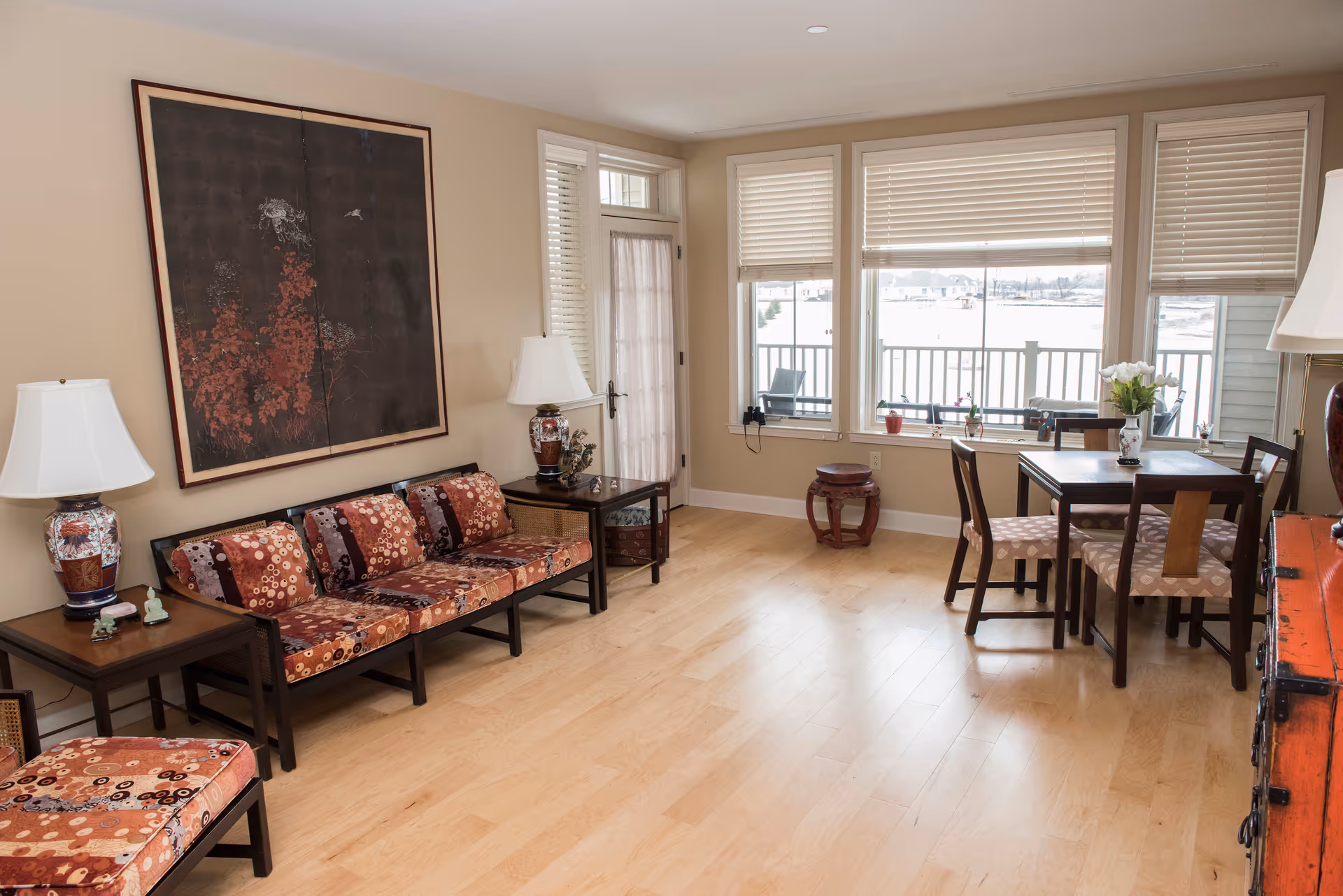 A bright living room with light wood flooring, a patterned sofa and chair, two side tables with decorative lamps, a large framed artwork on the wall, a small dining table with four chairs near large windows with blinds, and a door leading to an outdoor balcony.