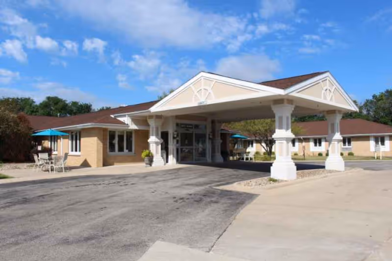 Exterior view of a single-story senior living facility building with a covered entrance supported by white columns. The building has beige brick walls and a brown roof. There are outdoor tables with blue umbrellas on the left side and trees in the background under a partly cloudy blue sky.