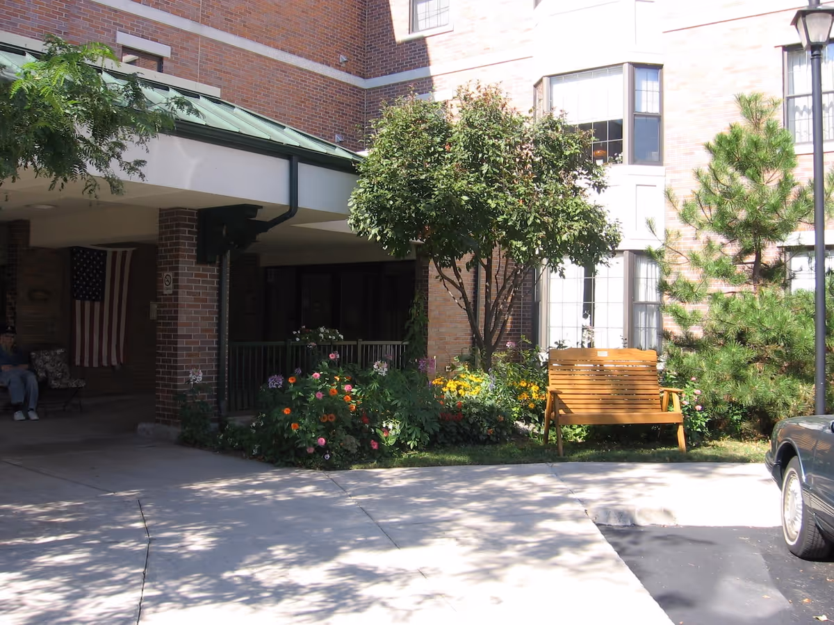 Front entrance of a brick senior living building with a covered driveway, flower beds, a wooden bench, and an American flag.