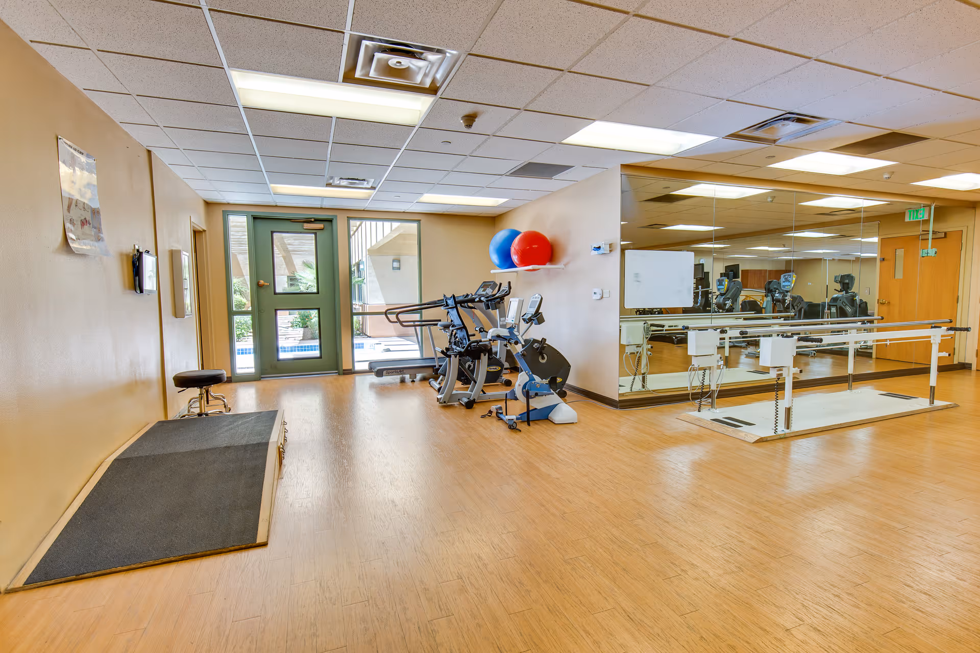 A spacious rehabilitation or physical therapy room with wooden flooring, a ramp, exercise equipment including stationary bikes, two large exercise balls on a wall shelf, and parallel bars for walking practice. The room has a large mirror on one wall and a glass door leading outside.