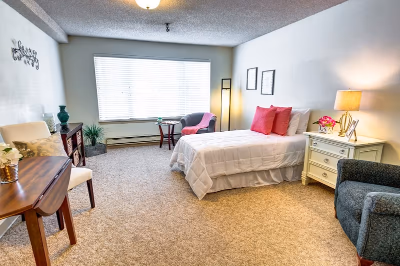 Bright furnished senior studio with a bed, nightstand, seating, and a large window.