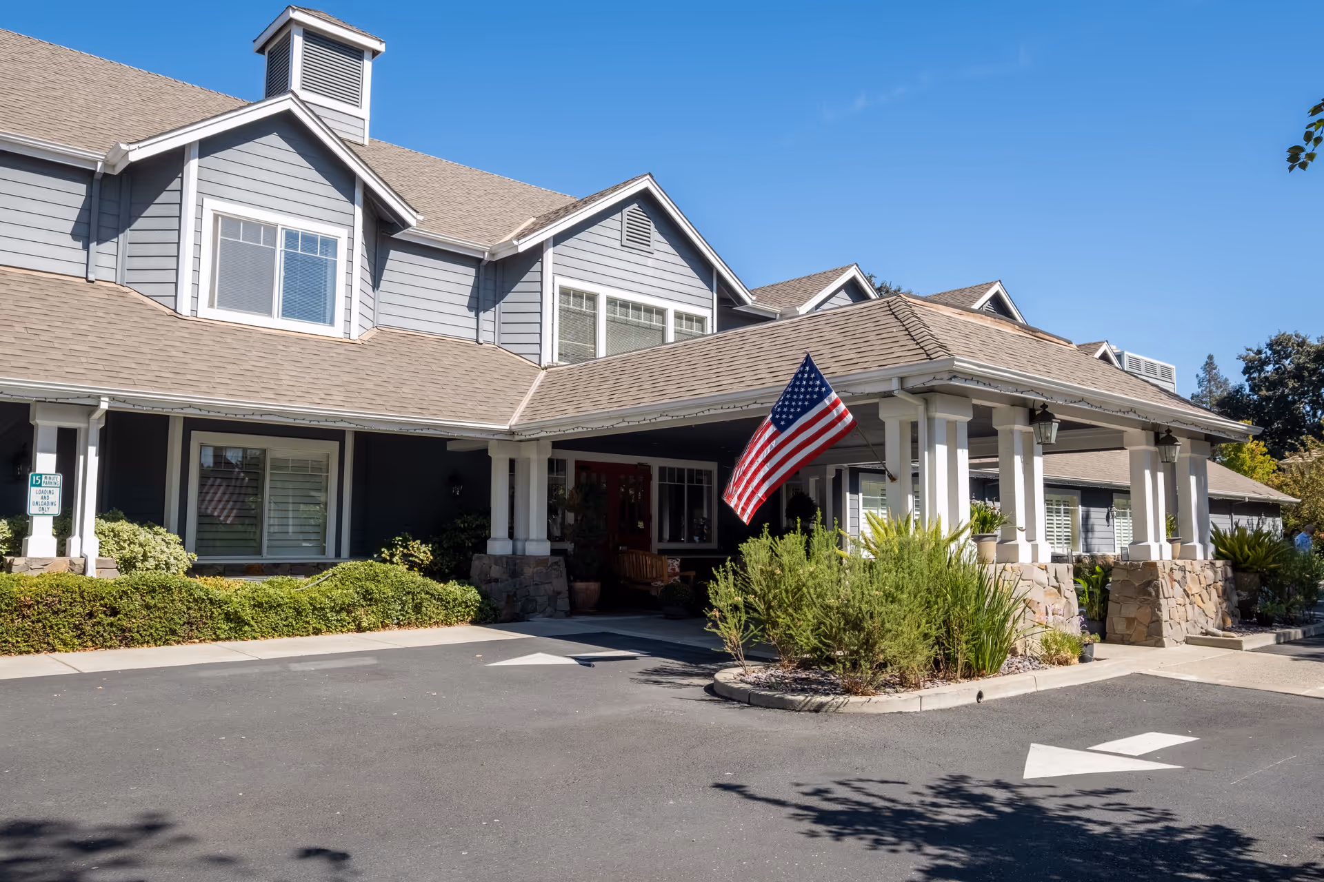 Exterior view of Aegis Living Pleasant Hill facility showing a large covered entrance with white pillars and stone bases, an American flag mounted near the entrance, and a clear blue sky overhead.