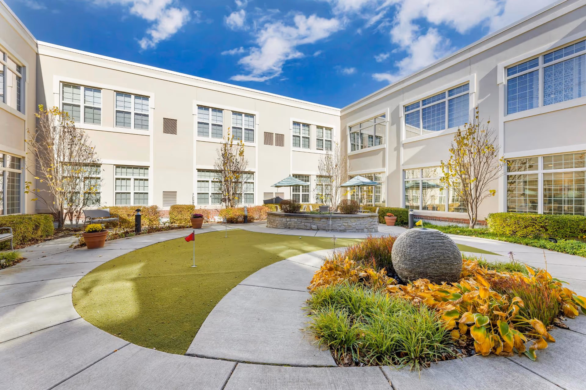 Outdoor courtyard area at North Shore Place featuring a small putting green with a red flag, surrounded by a concrete walkway. There are landscaped plants and a round stone water feature with yellow and green foliage. The courtyard is enclosed by a two-story building with large windows and a bright blue sky with some clouds overhead.