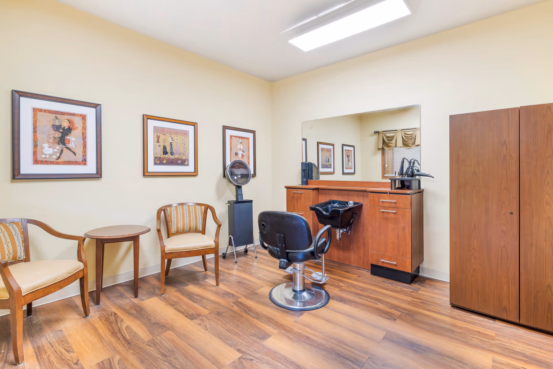 A small salon room with wooden flooring, two wooden chairs with cushioned seats and a small round table between them. There is a black salon chair in front of a wooden cabinet with a built-in sink and a large mirror above it. Three framed pictures hang on the wall, and a large wooden wardrobe stands to the right.