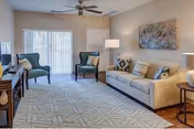 Bright living room with a sofa, two armchairs, patterned rug, side tables and lamps, ceiling fan, and sliding glass door with vertical blinds.