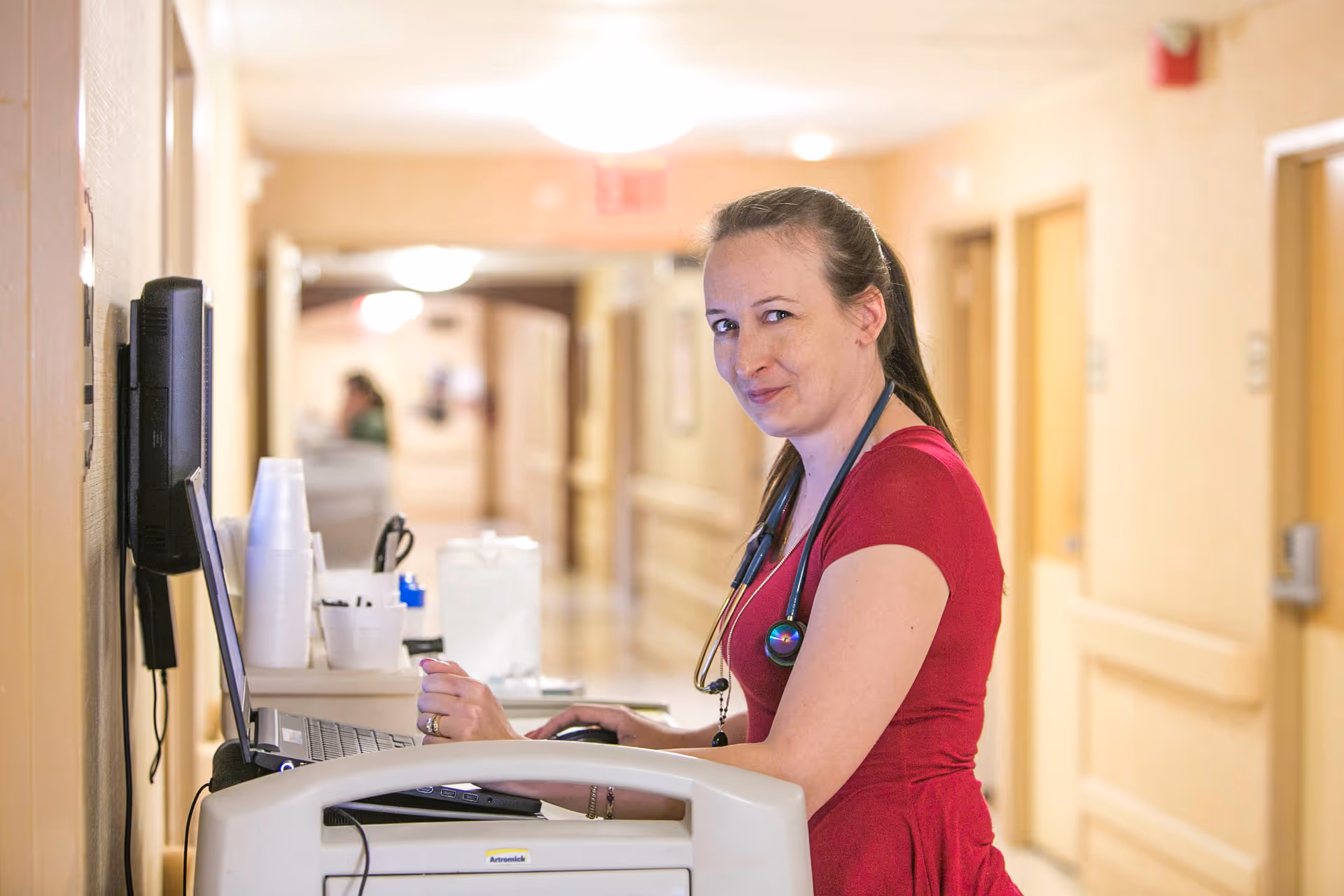 A female healthcare worker wearing a red shirt and a stethoscope around her neck is standing in a hallway, working on a laptop placed on a medical cart. The hallway has beige walls and doors on both sides, with another person blurred in the background.