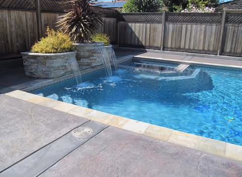 Backyard swimming pool with a built-in spa, stone waterfall planters, and surrounding concrete deck next to a wooden fence.