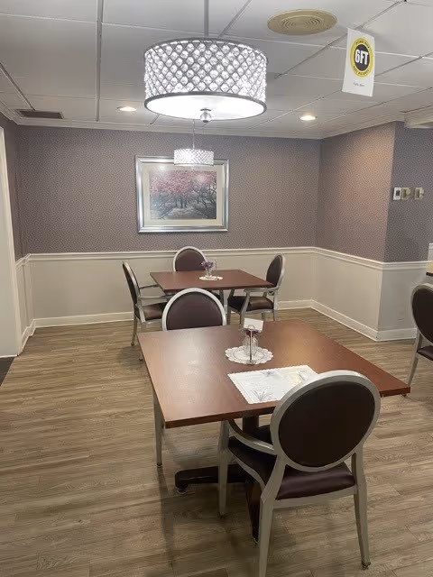 A small dining area with two wooden tables, each surrounded by four chairs with dark cushions. The room has light wood flooring, patterned wallpaper on the upper half of the walls, and white wainscoting on the lower half. Two modern ceiling lights hang above the tables, and a framed picture of pink trees is mounted on the wall. A sign indicating '6 FT' social distancing hangs from the ceiling.