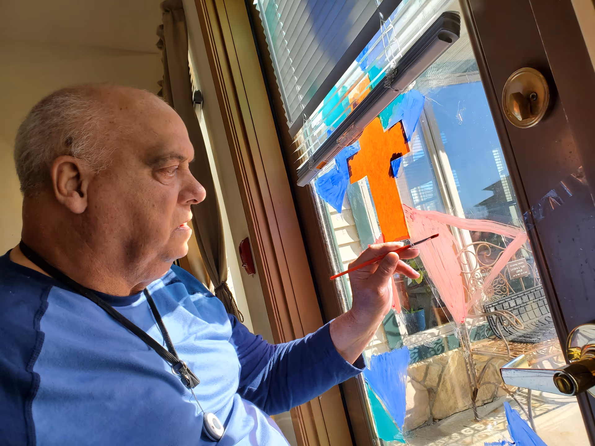 An elderly man wearing a blue shirt is painting colorful shapes, including a large orange cross, on a glass door with a paintbrush. Sunlight streams through the window, illuminating the scene and showing an outdoor patio area beyond the door.