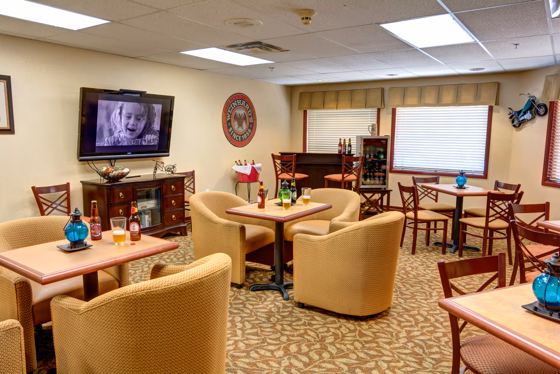A cozy common area with several tables and chairs arranged for socializing. There are cushioned armchairs and wooden chairs around tables with decorative blue glass lanterns and bottles of beverages. A flat-screen TV is mounted on the wall showing a black-and-white image of a child. The room has carpeted flooring with a patterned design, beige walls, and windows with blinds and valances. A small bar area with stools and a mini fridge stocked with drinks is visible in the background.
