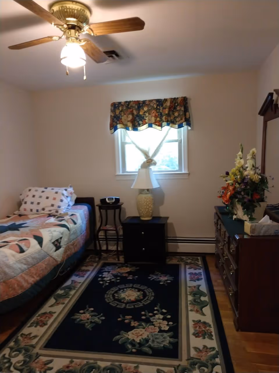 Small bedroom with a single bed, floral quilt, patterned rug, dresser with flowers, and a window with a valance.