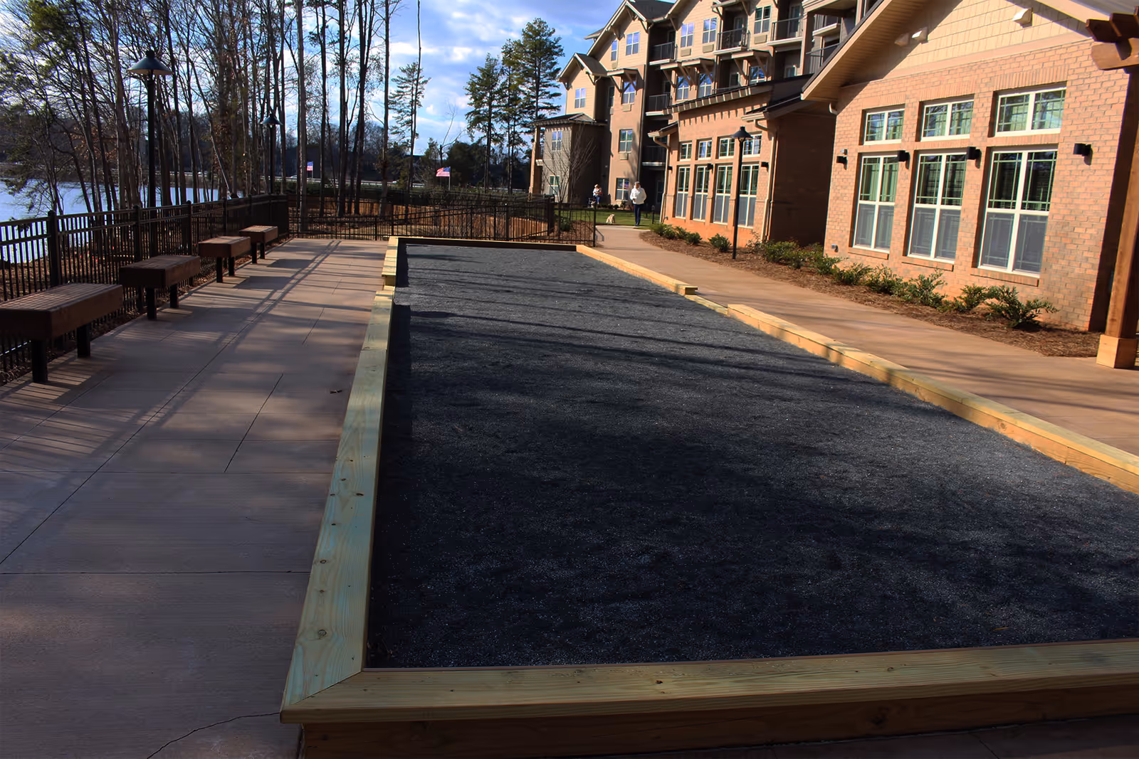 Outdoor courtyard showing a long gravel bocce court beside a multi-story brick senior living building with benches, trees, and a lake in the background.