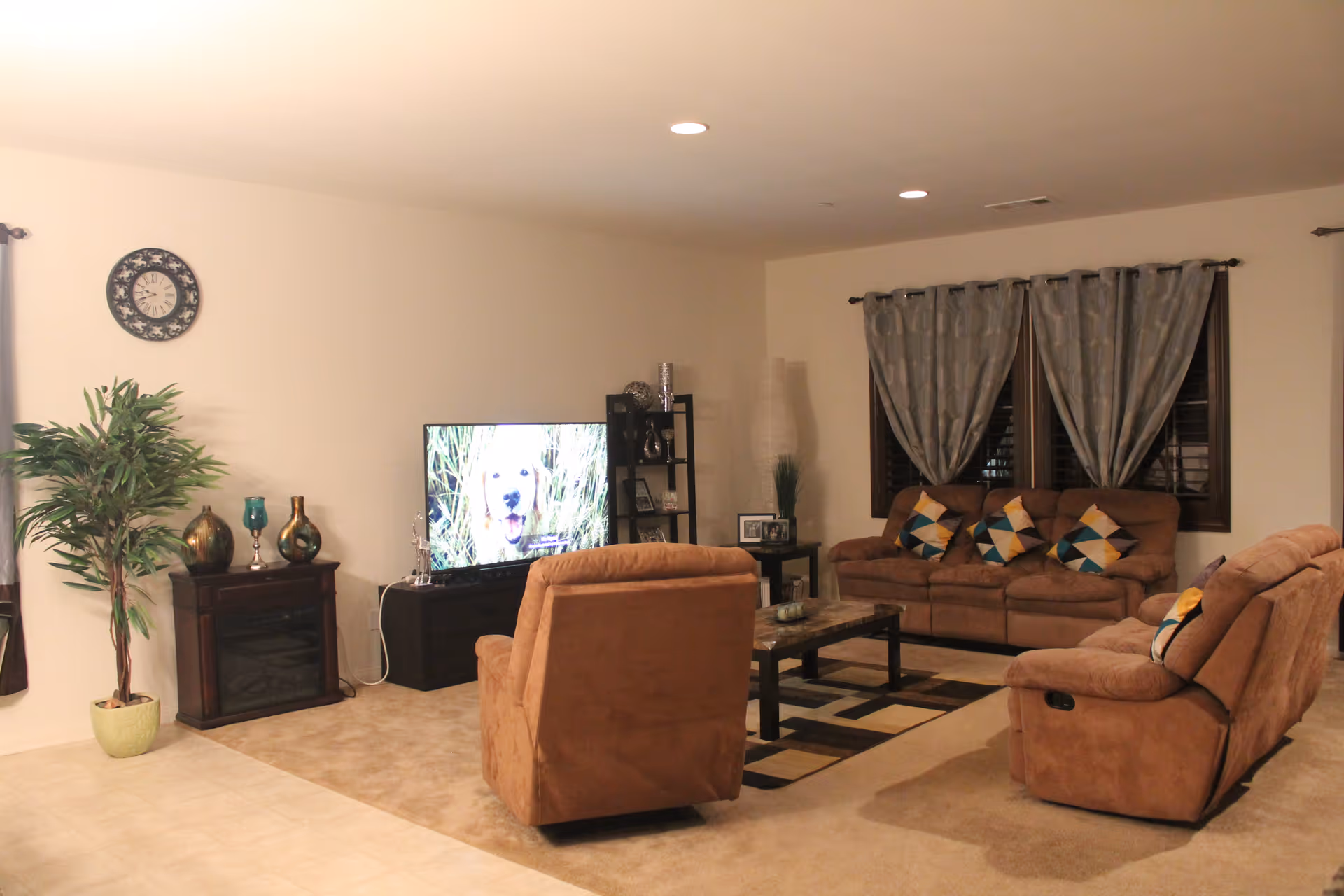 A cozy living room with beige walls and carpet. The room features a brown three-seater sofa and two matching recliners, all adorned with colorful geometric-patterned throw pillows. A rectangular coffee table sits on a patterned area rug in the center. A flat-screen TV displaying a dog is placed on a black TV stand, next to a dark wooden cabinet with decorative vases and a potted plant. Gray curtains cover the windows, and a round wall clock hangs on the wall.