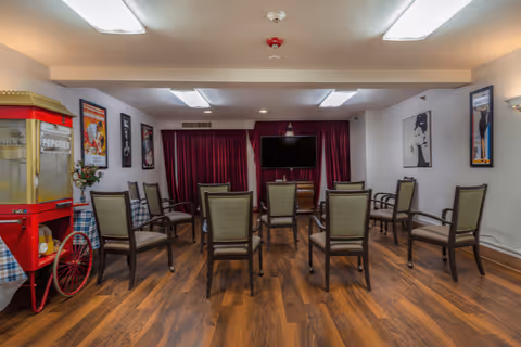 A room with multiple chairs arranged in rows facing a wall-mounted television. The room has wood flooring, white walls, and red curtains behind the TV. On the left side, there is a vintage-style popcorn machine and framed posters on the walls.